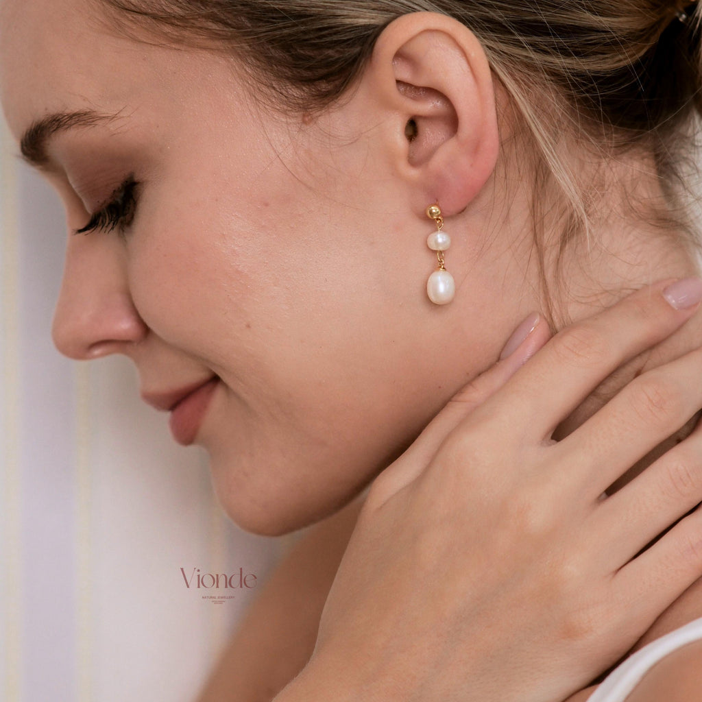 May include: Gold earrings featuring two white, pear-shaped pearls. The earrings are worn by a person with fair skin. The earrings have a simple, elegant design. The brand name "Vionde" is visible in the lower left corner.