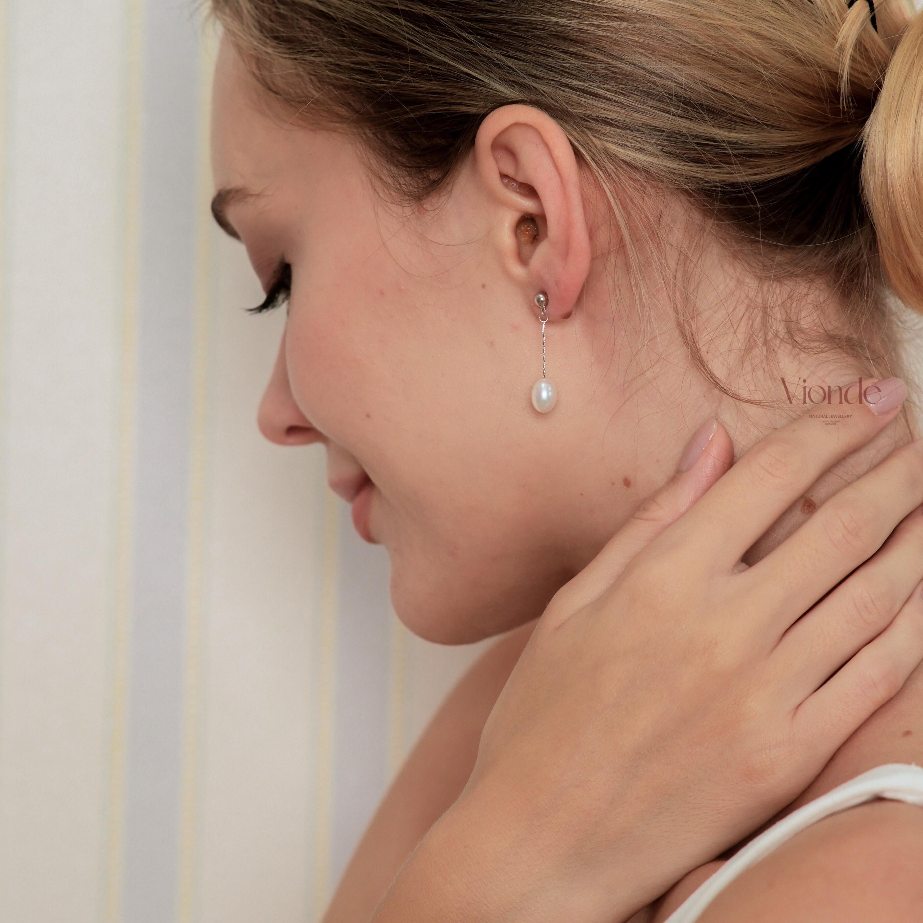 May include: A woman wearing a single pearl drop earring. The earring features a small stud with a delicate chain suspending a white, oval-shaped pearl. The background is a soft, blurred neutral color. The earring is a classic piece of jewellery.